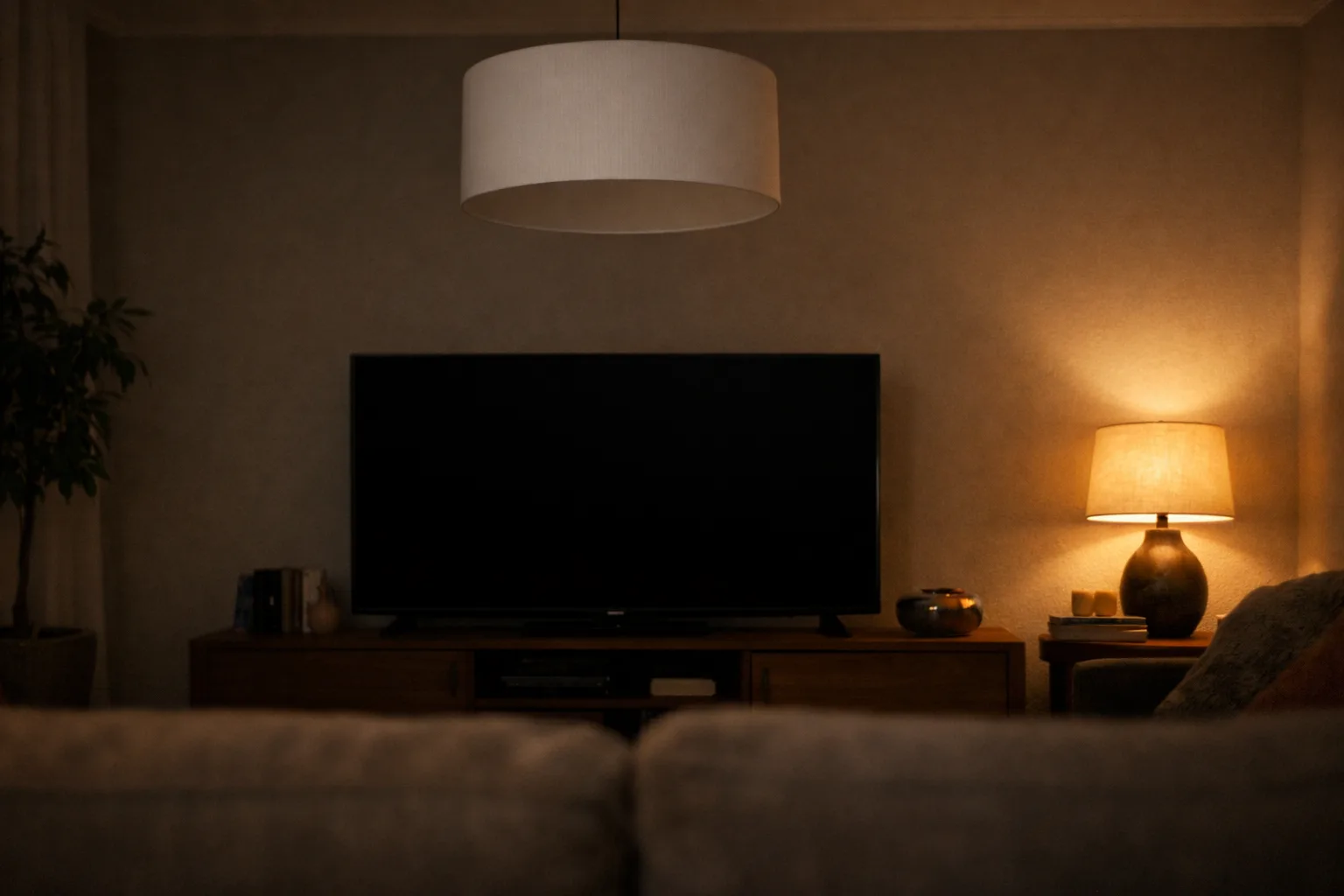 Dans un salon, une lampe d’appoint éclaire le mur à côté d’une télévision sombre, sous une suspension à abat-jour opaque.